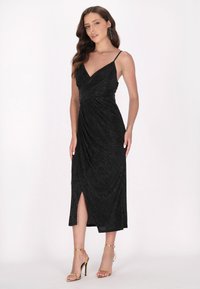 Schwarzes Kleid mit dünnen verstellbaren Trägern, V-Ausschnitt und seitlichem Schlitz. Aus strukturiertem Stoff mit drapiertem Design. Getragen mit goldfarbenen Absätzen.