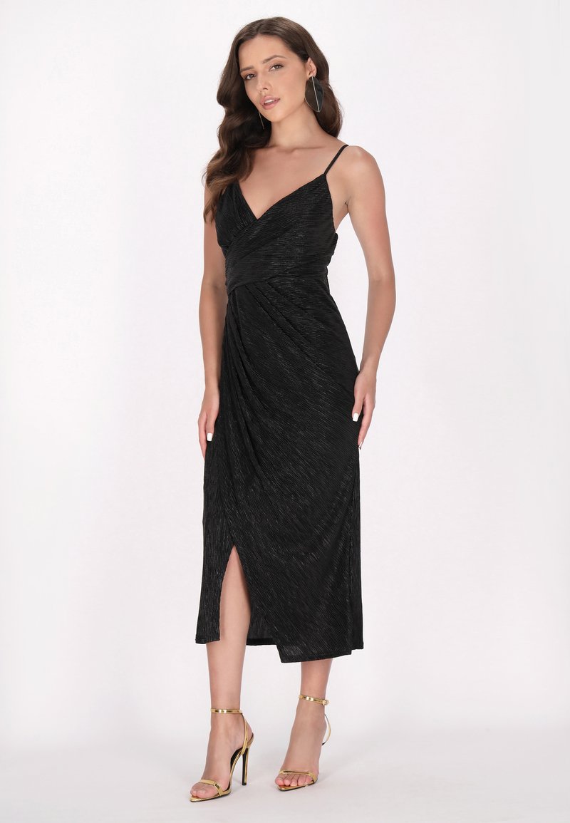 Schwarzes Kleid mit dünnen verstellbaren Trägern, V-Ausschnitt und seitlichem Schlitz. Aus strukturiertem Stoff mit drapiertem Design. Getragen mit goldfarbenen Absätzen.