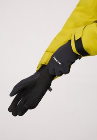 Gants noirs en matériau flexible, avec une prise texturée et une sangle de poignet réglable, associés à un manchon de veste jaune.