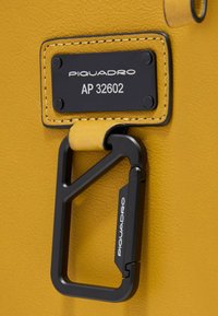 Gele lederen label met een zwarte metalen karabijnhaak, voorzien van het merknaam en modelnummer in zilverkleurige reliëfletters.