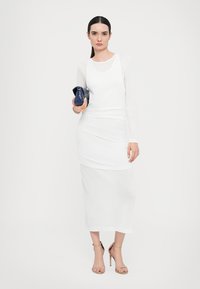 Witte lange mouwen top en een enkel-lange rok outfit, met een strakke, zachte textuur en ronde halslijn. Houdt een kleine blauwe clutch vast.