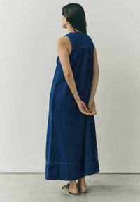 Rochie lungă din denim fără mâneci, de culoare albastru închis, cu buzunare laterale și o croială lejeră. Prezintă cusături vizibile de-a lungul cusăturilor și un tiv de lungime până la podea.
