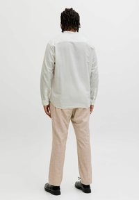 Personne aux cheveux courts et bouclés vue de dos, portant une chemise blanche à manches longues, un pantalon beige et des chaussures noires sur un fond uni.