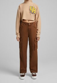 Beige kortärmad sweatshirt med logga; bruna cargobyxor med sidofickor; vita sneakers med mörka accenter; enkel grå bakgrund.