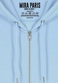 Felpa con zip di colore azzurro chiaro realizzata in cotone biologico. Presenta una zip in metallo, cordini e una morbida texture interna. Etichetta del marchio visibile all'interno.