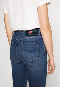 Tmavě modré džíny typu denim s přiléhavým střihem, s zelenou vyšitou nášivkou na zadní straně s červenými květinovými detaily a názvem značky.