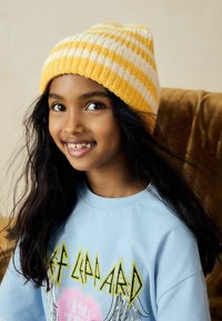 Bonnet tricoté rayé jaune et blanc ; cheveux longs et foncés ; t-shirt bleu clair avec design graphique ; souriant avec des dents visibles.