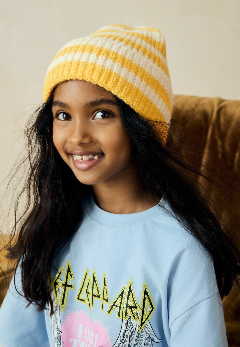 Bonnet tricoté rayé jaune et blanc ; cheveux longs et foncés ; t-shirt bleu clair avec design graphique ; souriant avec des dents visibles.