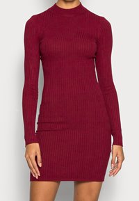 Abito a maglia a coste in bordeaux con collo alto e maniche lunghe, silhouette aderente e tessuto testurizzato che mette in risalto le righe verticali.