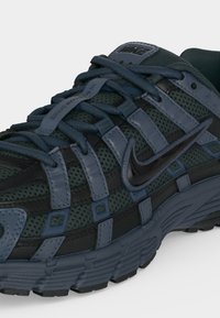 Σκούρο μπλε και μαύρο αθλητικό παπούτσι Nike με άνω μέρος από δίχτυ, δερμάτινες επικαλύψεις, χοντρή σόλα και κορδόνια δεμένα σε σταυρωτό μοτίβο.