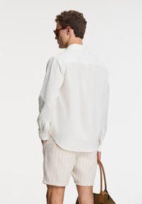Weißes Leinenhemd mit langen Ärmeln und Knopfmanschetten, kombiniert mit hell gestreiften Shorts. Das Model hält eine braune Tasche und trägt eine Sonnenbrille.