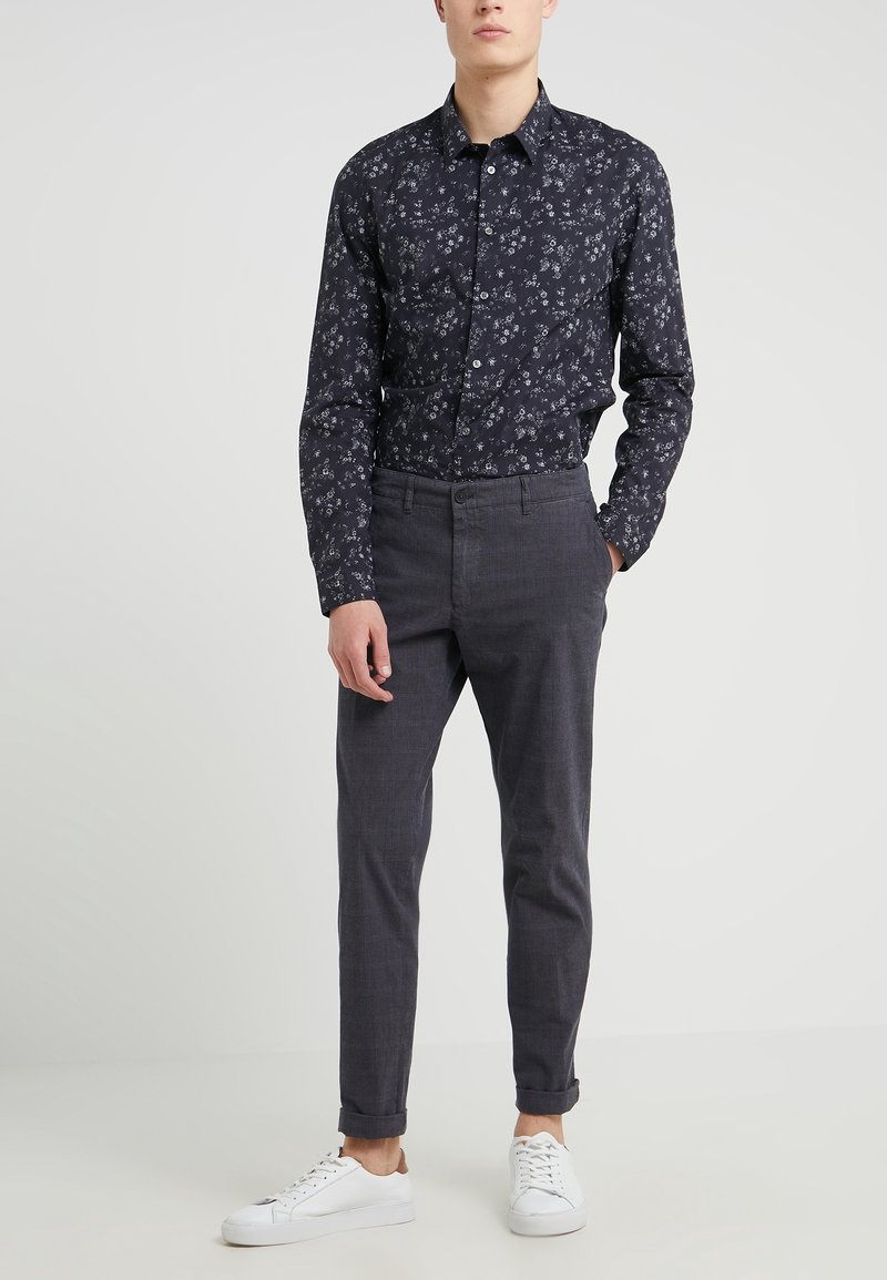 Homme portant une chemise noire à manches longues avec imprimé floral, un pantalon gris slim avec des revers et des baskets blanches, debout devant un fond uni.