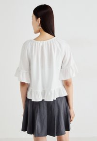 Blouse blanche à volants avec des manches courtes, présentant un col froncé et un ourlet en couches, associée à une jupe évasée gris foncé.