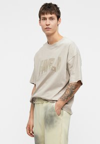 Tricou grafic bej cu inscripția "FAVELA" într-un print texturat, asociat cu pantaloni verzi deschis ce prezintă un model uzat, decolorat.