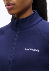 Κοντινό πλάνο ενός ατόμου που φοράει μπλε σκούρο μπουφάν Calvin Klein με φερμουάρ, με το λογότυπο στο στήθος και μέρος του κάτω μέρους του προσώπου του.