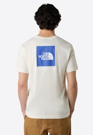 Cream-colored t-shirt featuring a blue logo square and text on the back. Cotton material with short sleeves and a crew neckline.