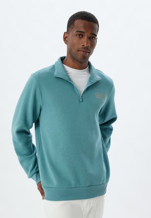 Lichtblauw pullover trui met een kwart-rits kraag, ribbels aan de mouwen en onderkant, en geborduurd logo-detail. Gemaakt van zacht, gestructureerd weefsel.