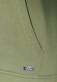 Olivenfarbener Sweatshirtstoff mit gerippter Textur, ausgestattet mit einem kleinen, silbernen Logotag und sichtbaren Nähdetails.