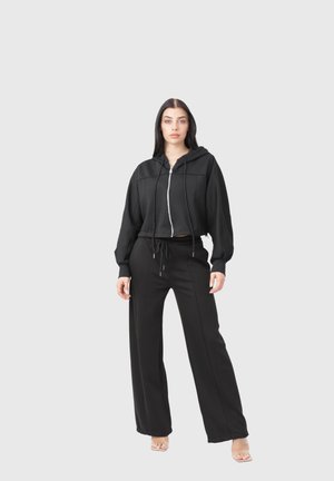 Vrouw draagt een zwart vest met rits en trekkoorden en wijde zwarte broek, staat blootsvoets op een effen lichtgrijze achtergrond.