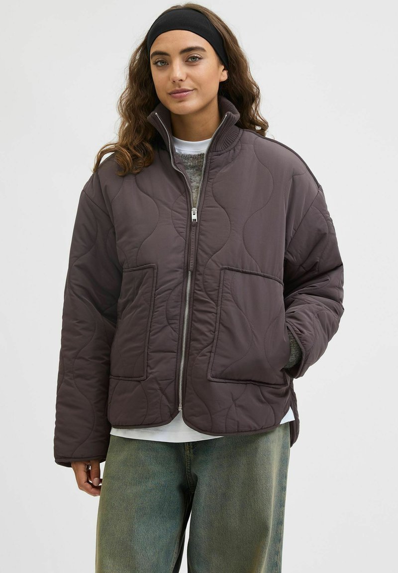 Veste matelassée rembourrée de couleur marron foncé, avec un col haut, une fermeture éclair à l'avant et deux poches avant. Portée par-dessus un haut blanc et un jean bleu clair.