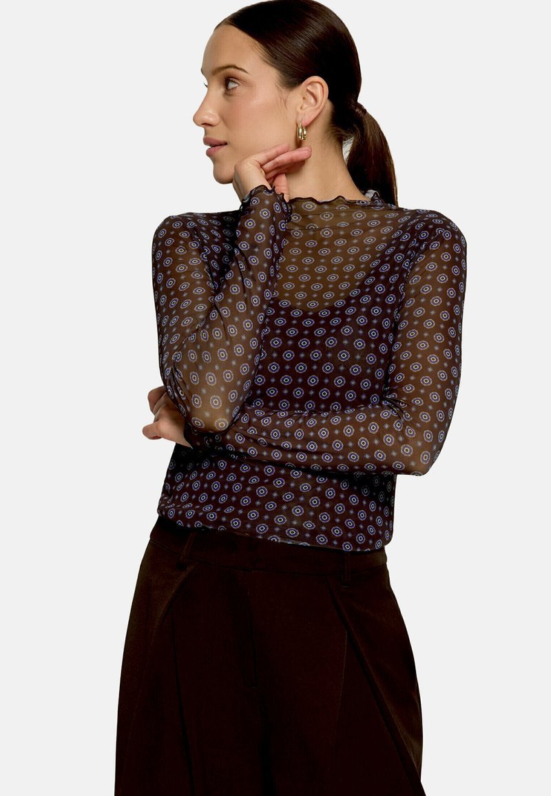 Femme aux cheveux foncés en queue de cheval portant un haut transparent à motifs à manches longues et un pantalon foncé, regardant sur le côté, la main près du visage.