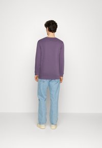 Suéter morado de manga larga con textura acanalada, escote redondo y hombros caídos. Combinado con jeans de mezclilla azul claro y zapatos beiges.