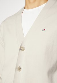 Cárdigan beige con cuello chal, con dos botones marrón claro y un pequeño parche con logo en la parte superior izquierda. Se lleva sobre una camiseta blanca.