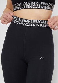 Schwarze Leggings mit einem breiten elastischen Bund, der den wiederholten Text "CALVIN KLEIN" zeigt. Der Stoff ist glatt, hat eine enganliegende Silhouette und dezente Logodetails.