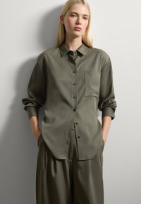 Blonde Frau in oversized olivgrünem Hemd mit Knöpfen und passenden weiten Hosen, steht mit Händen in den Taschen vor einer weißen Wand.