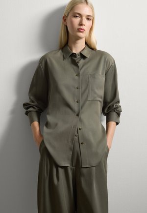 Donna bionda che indossa una camicia verde oliva oversize con bottoni e pantaloni larghi coordinati, in piedi con le mani nelle tasche contro un muro bianco.