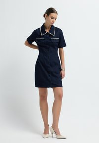 Dżinsowa sukienka z krótkim rękawem w ciemnoniebieskim kolorze, z dużym kołnierzem, dwiema kieszeniami na piersi i zapięciem na guziki, zestawiona z kremowymi butami na wysokim obcasie.