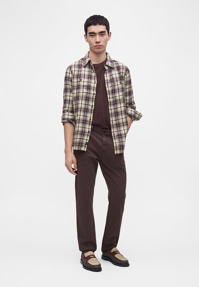 Jeune homme debout portant une chemise à carreaux beige et marron sur un t-shirt marron foncé, un pantalon marron foncé et des chaussures beiges et marron.