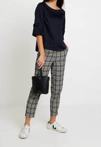 Marinblus med manschettärmar, rutiga byxor i svart och vitt samt vita sneakers med svarta detaljer. Håller en liten svart handväska.