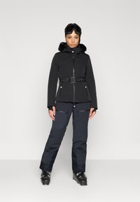 Μαύρο μπουφάν με μόνωση και κουκούλα από συνθετική γούνα, με φερμουάρ μπροστά και ζώνη με σχέδιο, σε συνδυασμό με σκούρα παντελόνια που έχουν πολλαπλές τσέπες.