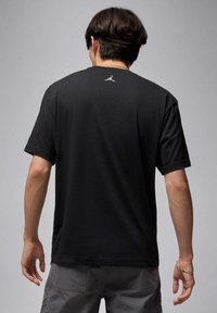 Czarny bawełniany t-shirt z krótkim rękawem, o luźnym kroju, z małym białym logo na górnej części pleców. Tekstura wydaje się gładka i miękka.