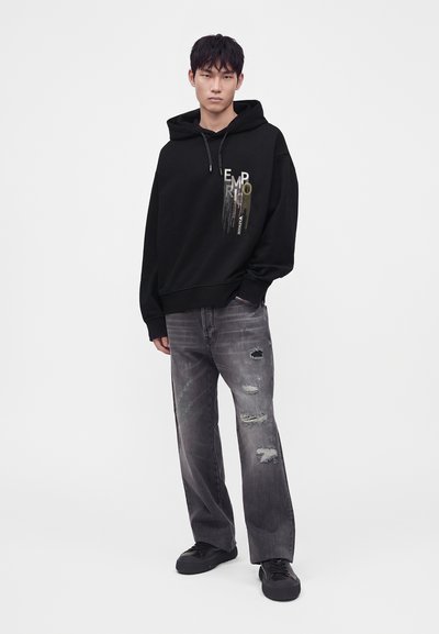 Jeune homme debout portant un sweat à capuche noir avec un motif, un jean gris usé et des baskets noires sur un fond blanc uni.