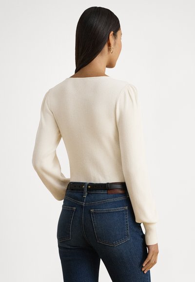Pull côtelé crème avec un large col et des manches longues bouffantes, associé à un jean bleu foncé et une ceinture noire avec un accent marron.