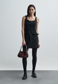 Schwarzes ärmelloses Kleid mit zwei dekorativen Taschen und Schleifenapplikationen. Kombiniert mit dunklen Strumpfhosen und einer kleinen bordeauxroten Handtasche.