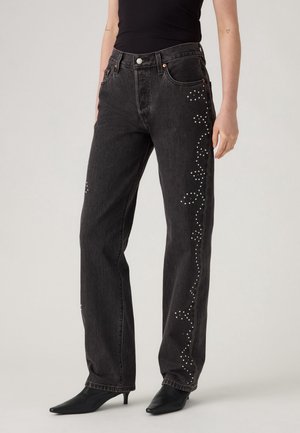 Femme portant un jean droit noir taille haute avec des motifs floraux blancs à pois le long des côtés, associé à des talons pointus noirs.