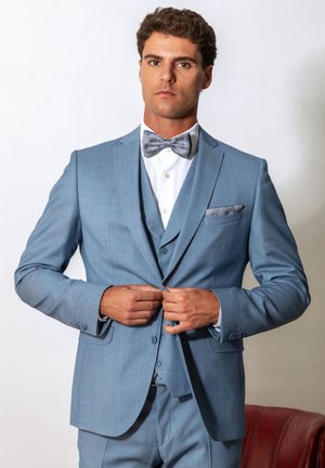 Mann, der einen Knopf an einer hellblauen Anzugjacke befestigt, trägt eine passende Weste, weißes Hemd und gemusterten Fliegenknoten, steht vor weißem Hintergrund.