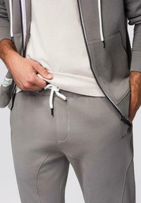 Homme portant un sweat à capuche gris zippé et un pantalon de survêtement assorti avec un cordon blanc, soulevant légèrement une chemise beige pour révéler la ceinture.