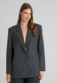 Grijze gestructureerde blazer met een doorgestoken ontwerp, diepe V-hals, twee knopen en twee voorzakken. Het materiaal heeft een subtiel patroon.