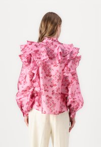Blusa rosa con stampa floreale, colletto e maniche arricciati, con una finitura satinata. Indossata con pantaloni color crema, dalla texture liscia.