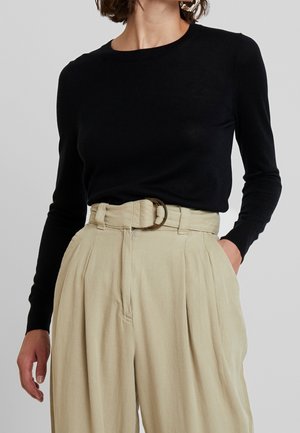 Kobieta w czarnej, długorękawowej bluzce wsuniętej w wysokowalne, beżowe plisowane spodnie z paskiem i okrągłą klamrą, ręką w kieszeni.