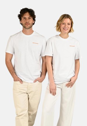 Homme et femme debout côte à côte portant des t-shirts blancs avec logo et pantalons de couleur claire sur un fond uni.