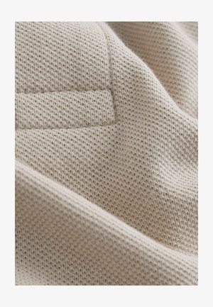 Bēša trikotāžas audums ar teksturētu rakstu, kurā redzama smalka režģa dizains. Mīksta, nedaudz pūkaina tekstūra ar locījumu malā.