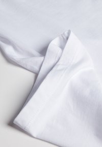 Camiseta blanca de algodón con una textura suave y una manga doblada. Presenta un diseño simple y limpio con costuras prolijas a lo largo de los bordes.
