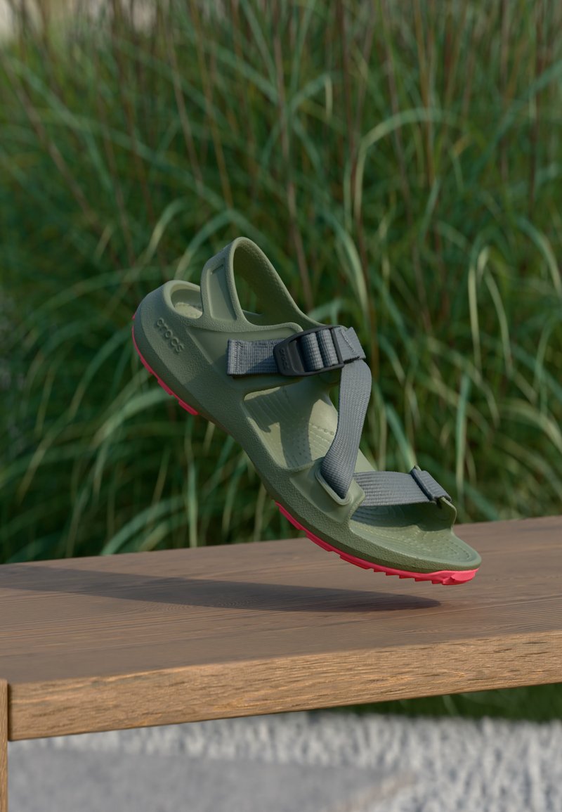 Sandale verte et rose avec des brides grises ajustables semble suspendue au-dessus d’un banc en bois en extérieur avec un arrière-plan d’herbes hautes.