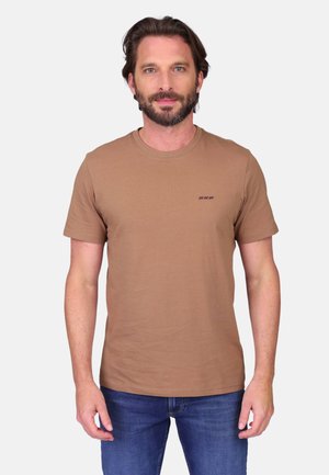 Homme barbu portant un T-shirt marron uni à manches courtes et un jean bleu, debout devant un fond gris clair.