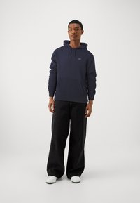 Mørkeblå hættetrøje med frontlommer, snoretræk i hætten og logo detaljer, parret med brede sorte bukser og hvide sneakers.